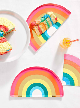 A vibrant rainbow-themed party setup featuring colorful paper plates, a multi-layered rainbow cake with a slice removed, and a swirly rainbow lollipop on a white background.