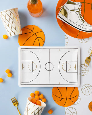 Flat lay of a basketball-themed party with snacks, a basketball court serving tray, decorative plates, and cups, with a playful sneaker illustration, set against a light blue backdrop.