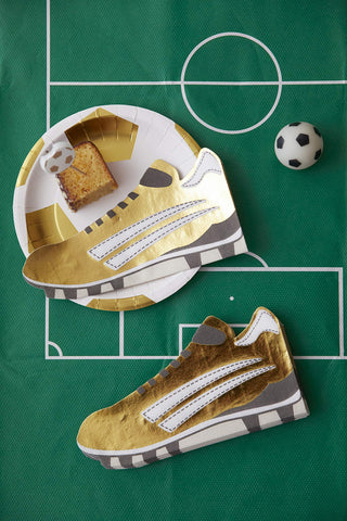 A creatively laid sports-themed tabletop featuring a football field tablecloth, a plate shaped like a sneaker with a piece of cake on top, and a matching sneaker alongside a miniature soccer ball.