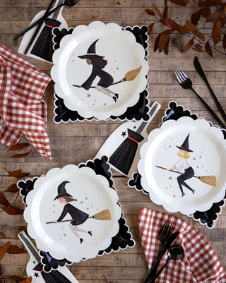 A festive Halloween party supplies table setting with My Mind's Eye Witches Paper Plate Set, black utensils, and red and white gingham napkins, arranged on a wooden surface with autumn leaves.