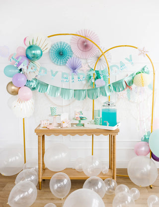 Colorful birthday party setup with a decorated table under a mylar fringe banner, featuring a blue punch bowl and a Mermaid Happy Birthday Banner Set from My Mind's Eye.