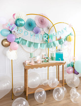A festive birthday party setup with a table displaying treats, balloons, and a "Mermaid Happy Birthday Banner Set" in soft pastel tones by My Mind’s Eye.