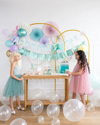 Two young girls at a festive birthday party, standing beside a table with cake and balloons, decorated with pastel streamers and a My Mind's Eye Mermaid Happy Birthday Banner Set.