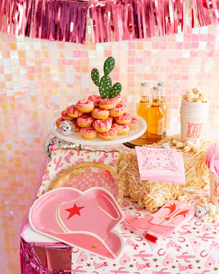 A vibrant party table with pink macarons on a cake stand, decorative cactus, themed beverages, and a My Mind's Eye cowgirl pattern paper plate, set against a pink tinsel and tiled backdrop.