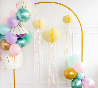 Colorful party balloons and My Mind's Eye Honeycomb Jellyfish Set hanging from a gold arch, with a white wall in the background.
