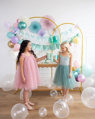 Two girls at a birthday party, one handing a My Mind's Eye Honeycomb Jellyfish Set to the other, surrounded by colorful balloons and under the sea decor.