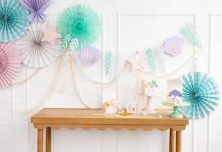 A festive party table with an under the sea theme, decorated with pastel-colored paper fans, seashells, and a net, featuring cakes and starfish-shaped treats on stands next to the Mermaid Jumbo Banner from My Mind's Eye.