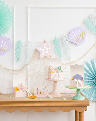 A festive party table decorated with pastel-colored seashell and star decorations, a Mermaid Jumbo Banner backdrop from My Mind's Eye, and various beach-themed items.