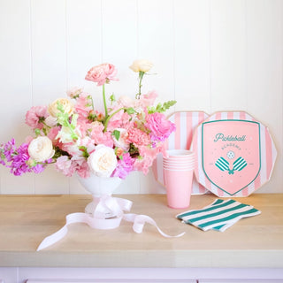 A vibrant arrangement of pink flowers in a white vase, accompanied by pink cups and Le Pickleball Small Plates made from eco-conscious materials by Bonjour Fête, set against a wooden surface with a pickleball paddle leaning on.