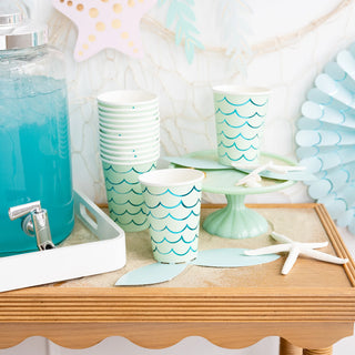 A beach-themed party setup with a blue beverage in a dispenser, My Mind's Eye Mermaid Tail Paper Party Cups on a tray, and decorative starfish, all against a backdrop of pale blue balloons.