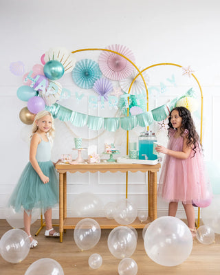 Two young girls in pastel tulle dresses at an under-the-sea-themed birthday party table decorated with aqua foil balloons, paper fans, and a My Mind's Eye Mermaid Tail Paper Party Cups drink dispenser.