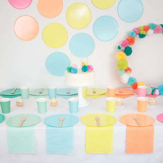 A pastel-colored birthday party setup with a white frosted cake at the center, surrounded by Loop by Frankie Aqua Paper Plates, cups, and a pompom garland on a white backdrop.