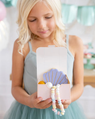Girl in a blue dress holding a My Mind's Eye Mermaid Treasure Treat Box Set with a decorative cookie, standing in a room with pastel balloons and streamers.