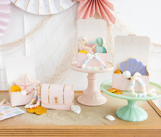 A display of children's under the sea-themed party decorations and accessories, including My Mind’s Eye Mermaid Treasure treat box sets, bracelets, and hair accessories on pastel-colored stands.