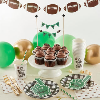 A festive table setup for a football-themed party, featuring chocolate cupcakes, decorative balloons, paper tableware, and cups with playful sports slogans, accompanied by Slant's Football Party in a Box - Tailgate decorations in gold and green.