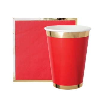 A Posh Ruby Kiss cup from Jollity & Co with a golden rim juxtaposed against a textured red background partially overlapped by a gold-splattered beige piece, creating a festive and luxurious party theme.