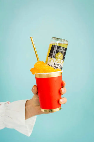 A person holds a Posh Ruby Kiss Cup from Jollity & Co, topped with a frozen orange slush and an upturned soda can, with a golden straw for sipping— a playful and vibrant addition to our luxe tableware collection.