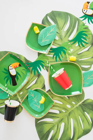 Colorful tropical-themed party table setting with green leaf-shaped plates, Jollity & Co gold foil Posh Ruby Kiss Cups, and paper toucan decorations on a white background.