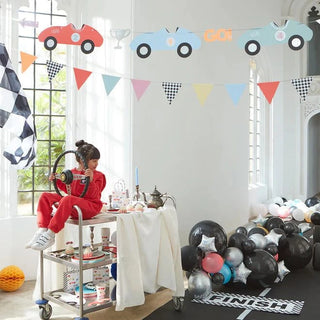 A child in a red outfit sits pensively at a race car-themed party, complete with Meri Meri's Vintage Race Car Garland, a checkered flag, and a table laden with treats amidst a balloon-adorned.