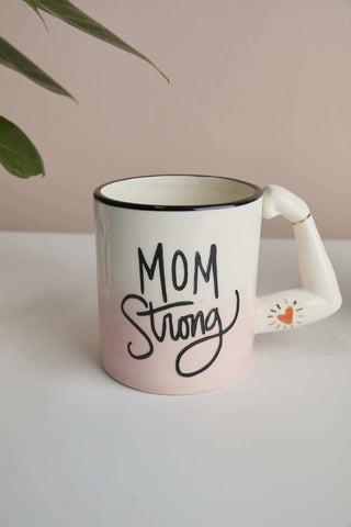 A pastel pink and white Accent Decor ceramic mug with "Mom Strong" written on it, placed against a taupe backdrop with a green plant leaf partially visible in the upper left corner. Visible handcrafted imperfections add to its charm.