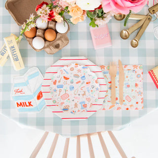 Table with a checkered tablecloth, floral centerpiece, eggs, butter, milk carton, sugar, and pastel-colored plates and cutlery.