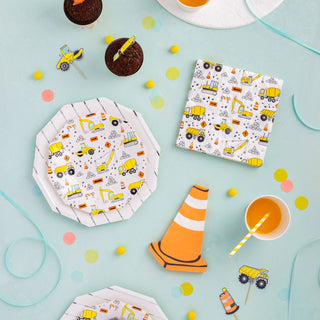 A cheerful construction-themed party setting with coordinating tableware, featuring playful designs of diggers and trucks, complemented by a scattering of colorful confetti and a striped paper straw, against a pastel blue backdrop.