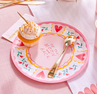 A sweet vanilla cupcake with white frosting and sprinkles rests on a heart-themed paper plate with "to the moon and back" text, alongside a golden spoon, on a pink tablecloth.