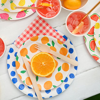 A colorful summer-themed picnic setting with fruit-patterned disposable tableware, a fresh orange slice on a plate, and refreshing drinks with orange slices and straws.