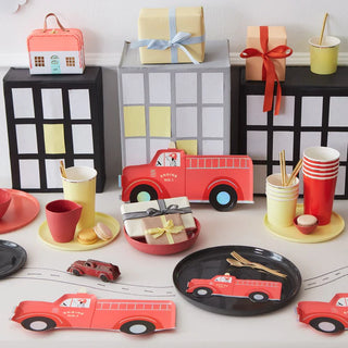 A themed party table setup with a firetruck motif, featuring coordinating tableware, decorations, and gifts, arranged against a backdrop of handcrafted buildings.