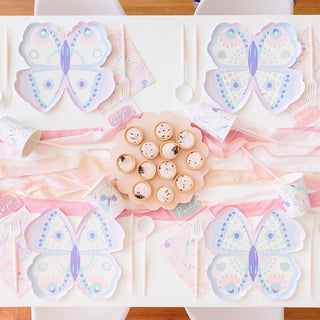 A pastel-themed party table is set with butterfly-shaped plates, matching cups, and party favors; a platter of cupcakes sits at the center, adorned with decorative toppers.
