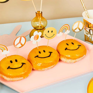 Delicious glazed doughnuts with cheerful smiley faces on a pastel pink plate, accompanied by cute, themed toppers featuring rainbows, suns, and peace signs, creating a vibrant, happy-go-lucky party atmosphere.