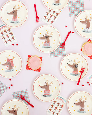 A festive table setting featuring plates adorned with whimsical reindeer illustrations, complemented by checkered napkins, red cutlery, and holiday-themed decorations, all laid out on a soft pink background.