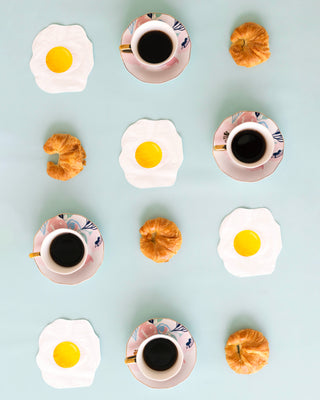 A flat lay image showing a pattern of three coffee cups, four croissants, and five fried egg coasters on a light blue background.