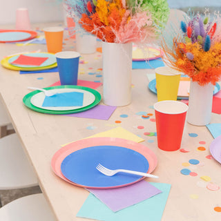 A vibrant party table set with colorful disposable plates, cups, and napkins, accented with bright confetti and whimsical floral centerpieces, ready for a cheerful celebration.