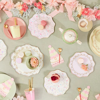 A pastel-themed tea party setup with floral-patterned porcelain, delicate macarons, and hints of gold on a soft green fabric with decorative flowers.