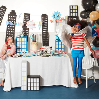 Two children at a superhero-themed party, one standing on a chair striking a pose with cape and mask, the other gazing upwards, with a table adorned with comic book-style decorations and snacks, surrounded by cityscape cutouts and balloons.