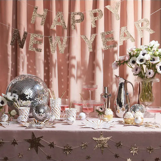 A festive new year celebration setup featuring a "happy new year" banner, disco balls, decorative stars, and a table adorned with flowers and partyware bathed in a warm, rosy glow.