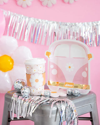 A pink-themed party setup with a floral-decorated cup, a pink van-shaped sign, small disco balls, and silver tinsel decorations on a gray stool. Pink backdrop with silver fringe and daisy decorations.