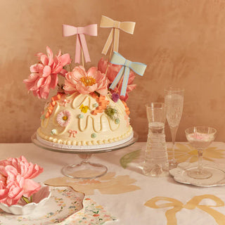 A pastel-themed celebration with an elegantly decorated cake adorned with pink flowers and delicate icing, flanked by chic glasses and complemented by floral accents on a warmly lit backdrop.