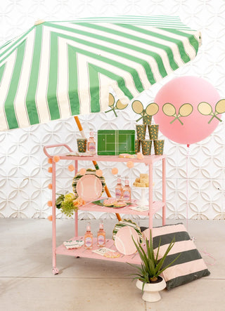 Pink bar cart with drinks and cups under a green and white striped umbrella. Decor includes tennis-themed balloons, plates, pillows, and a small plant, set against a white textured wall.
