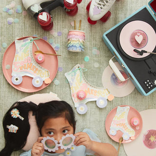 A child lies on the floor amid a colorful setup with retro roller skates, vinyl records, pink-themed party plates, and decorations suggesting a fun, vintage-themed celebration.