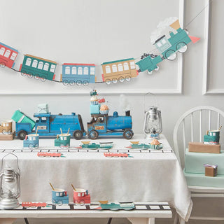 A birthday party table is festively decorated with a transportation theme, featuring a colorful paper train banner, toy vehicles, and coordinated tableware and centerpieces.