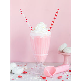 A creamy pink milkshake topped with whipped cream in a classic glass with two decorative straws, surrounded by sweet confections and heart-shaped sprinkles on a pastel backdrop.