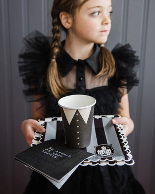 A contemplative young girl in a black tulle dress holds a stylish paper cup and a themed napkin, possibly at a chic party or event.