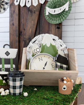 A rustic-themed party setup featuring a wooden crate with paper decorations resembling a campsite, including a clock designed like a mountain, a cup with popcorn, and a snack box resembling a backpack, on a faux grass surface.