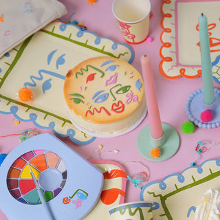 A decorated table with a round cake featuring colorful face designs, surrounded by festive plates, candles, a paint set, and pom-poms on a pink surface.