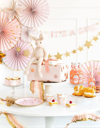 A pastel-themed baby shower setup with a "it's a girl" sign, a cake topped with a plush kangaroo, coordinating paper fans, star decorations, and assorted sweets on a white table.