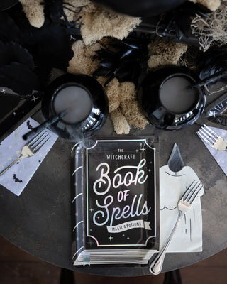 A mystical tablescape features a witchcraft spellbook centerpiece, flanked by black plates, silverware, and wine glasses, evoking a dark, magical dinner theme.