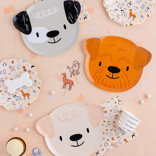 Assorted dog-themed party tableware, including plates with cute canine illustrations in soft tones, displayed on a peach-colored background with a sprinkling of confetti.