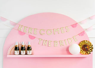 A festive wedding celebration decor with a pink backdrop featuring a "here comes the bride" banner, along with champagne bottles, pink rosettes, a white ornament, and a paper fan on a shelf.